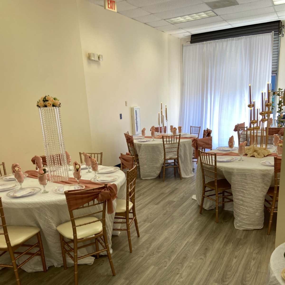 A banquet hall arranged with round tables covered in white tablecloths, adorned with decorative centerpieces and chairs set with light brown cushions.