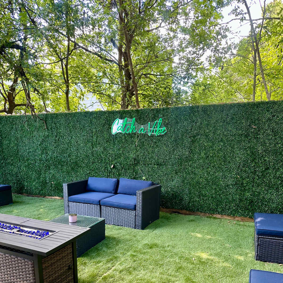 A seating area with dark wicker furniture is set against a green hedge featuring a neon sign that says "Catch a Vibe."