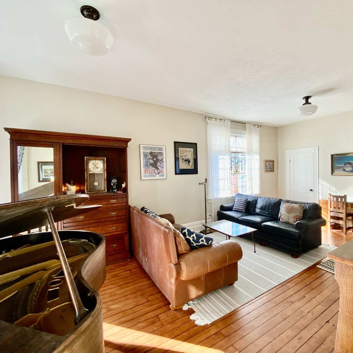 A bright living room featuring a piano, two sofas, a coffee table, and a wooden cabinet against a wall with several windows.