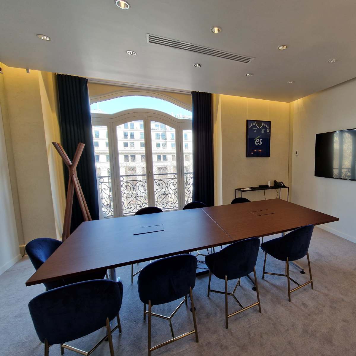 A modern conference room features a large table, plush chairs, a wall-mounted display, and a balcony with a view.