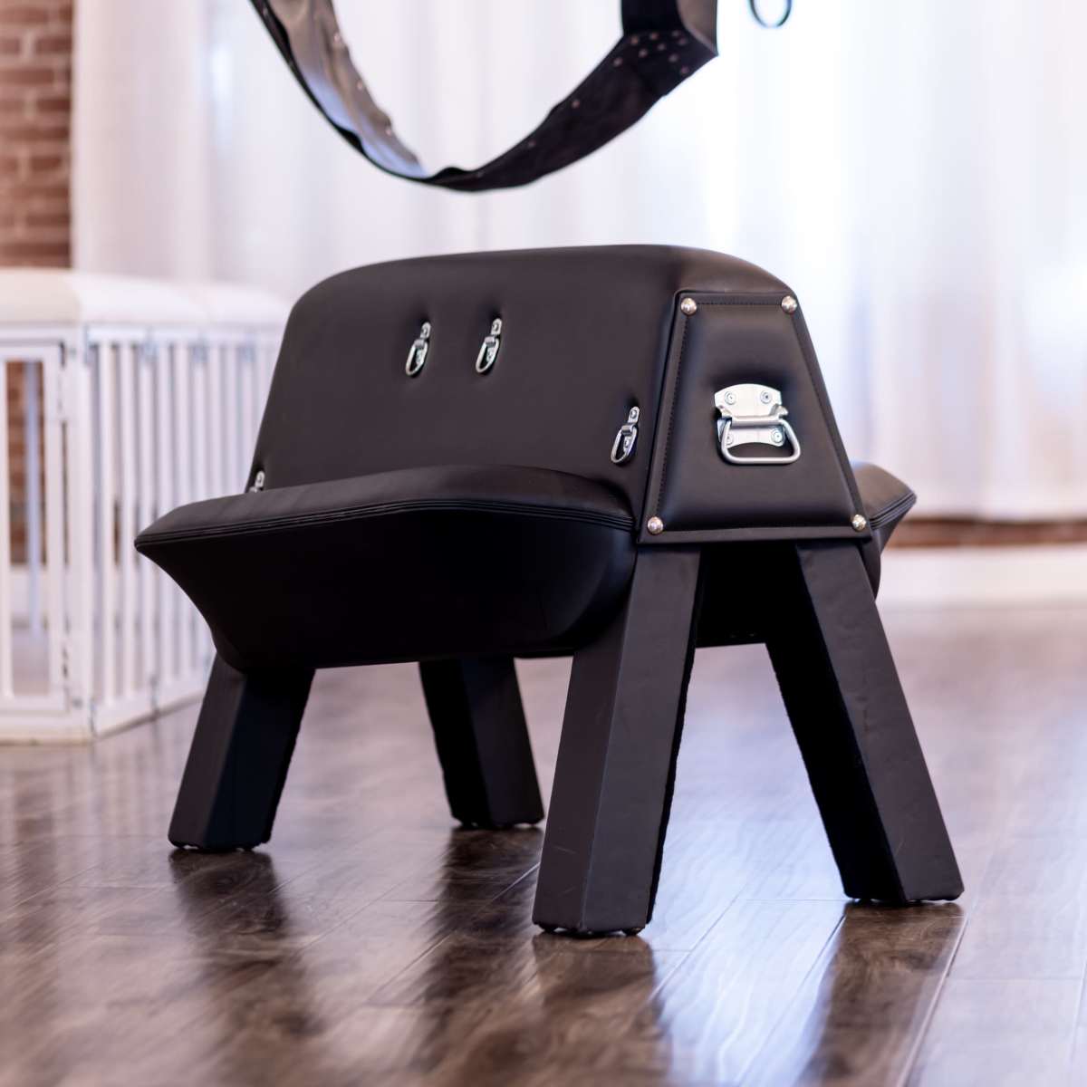 A black leather bench with metallic fasteners and a sturdy, angular frame, set against a backdrop of wooden flooring and bright drapes.