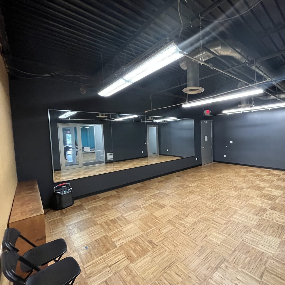 An empty studio room with dark walls, a large mirror, and wooden flooring, featuring two chairs and a trash can.