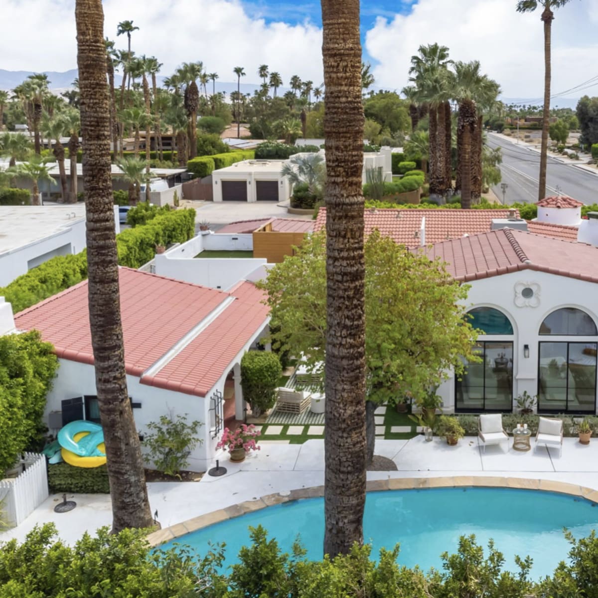 A single-story house with a red tile roof is surrounded by palm trees and features a pool with lounge chairs in the backyard.