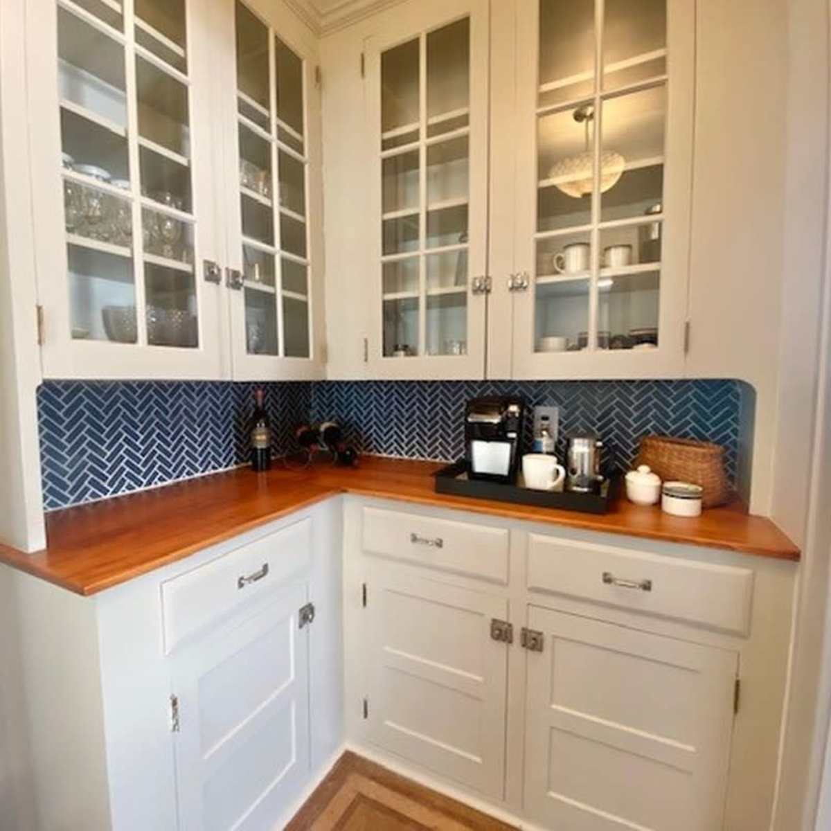 A kitchen nook featuring white cabinets with glass doors, a wooden countertop, and a coffee station against a blue herringbone backsplash.