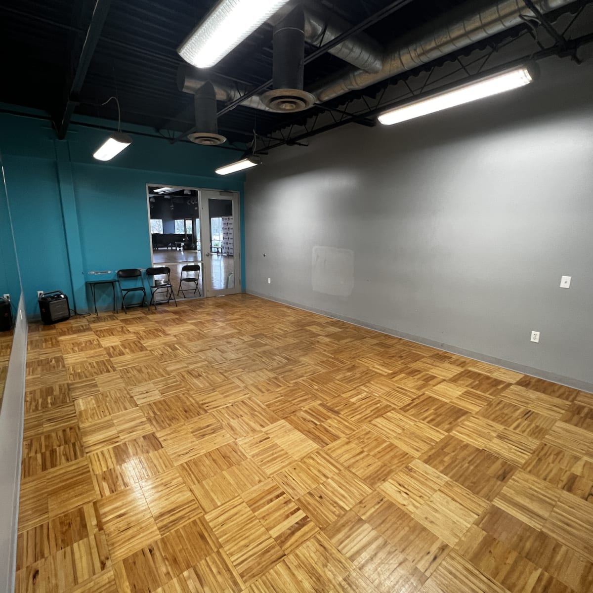 An empty room with wooden flooring, a mirrored wall on one side, and teal-colored and gray walls.