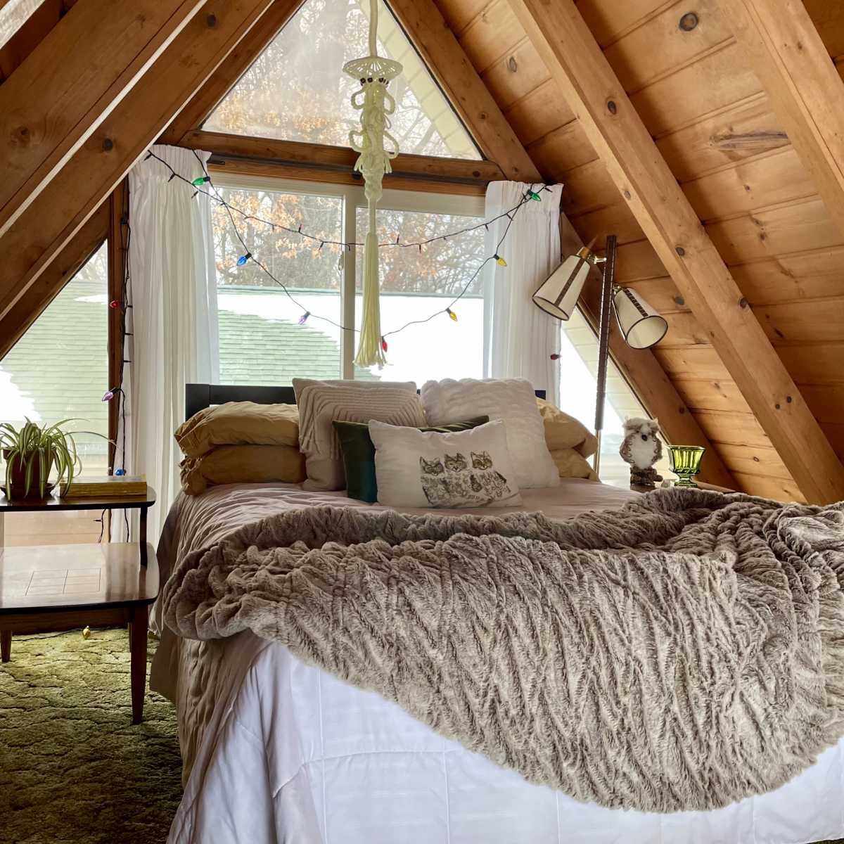 The bedroom features an A-frame design with a large window, a cozy bed adorned with decorative pillows and a knit blanket, and wooden walls.