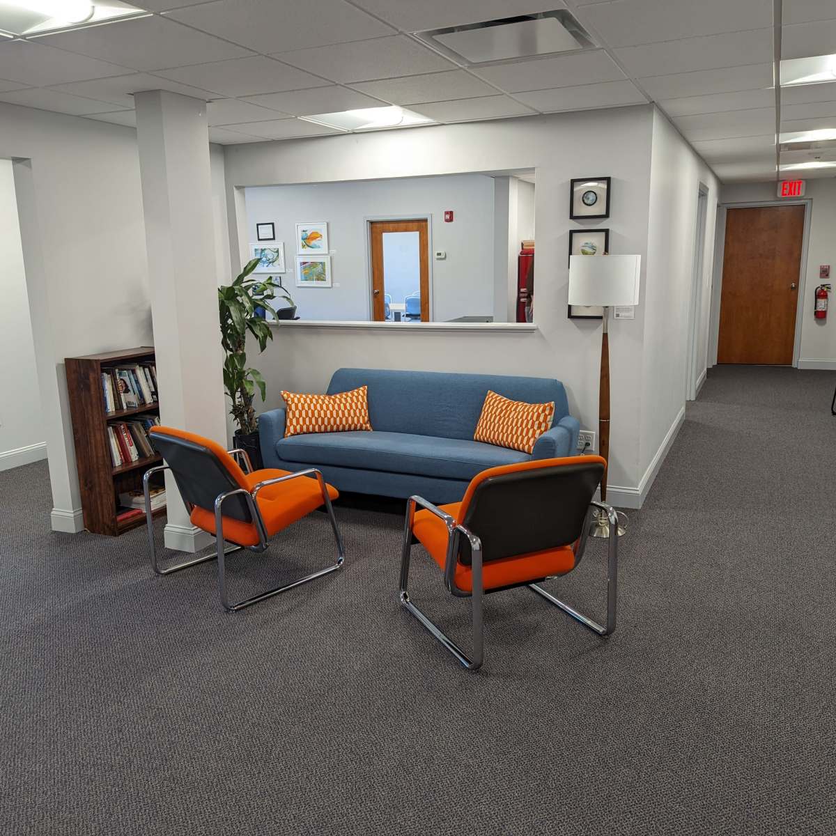 A modern office waiting area features a blue sofa with orange accent pillows and two orange-accented chairs, alongside a plant and bookshelves.