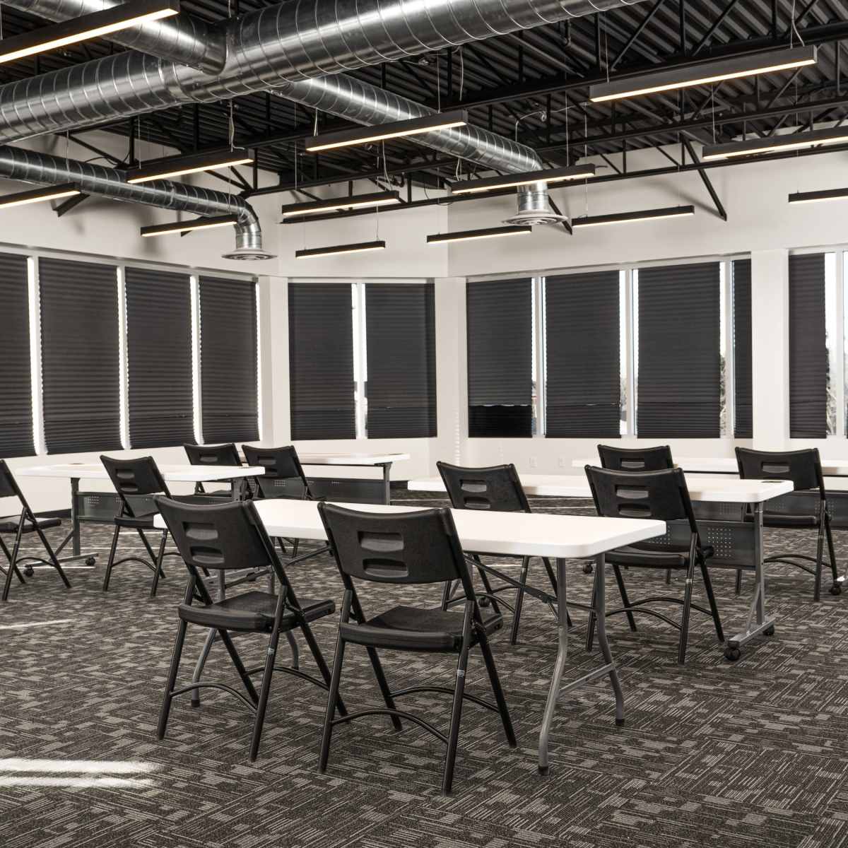 A classroom features several rows of white tables and black chairs arranged on a carpeted floor, with large windows covered by retractable blinds.