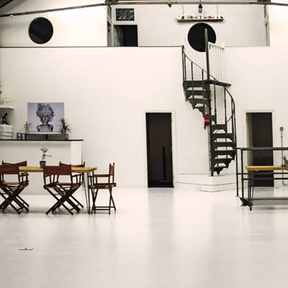 A spacious, minimalist studio with white walls, a spiral staircase, and various furniture arrangements including tables and stools.
