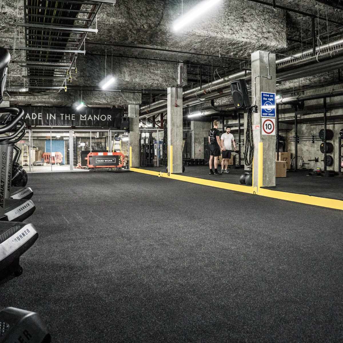 A spacious underground gym features various exercise equipment, including treadmills and weights, with two individuals engaging in a workout.