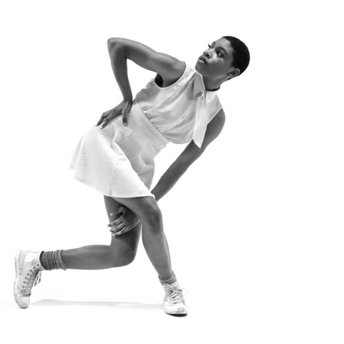 A person in a white dress and sneakers poses with one hand on their hip and the other on their knee against a plain white background.