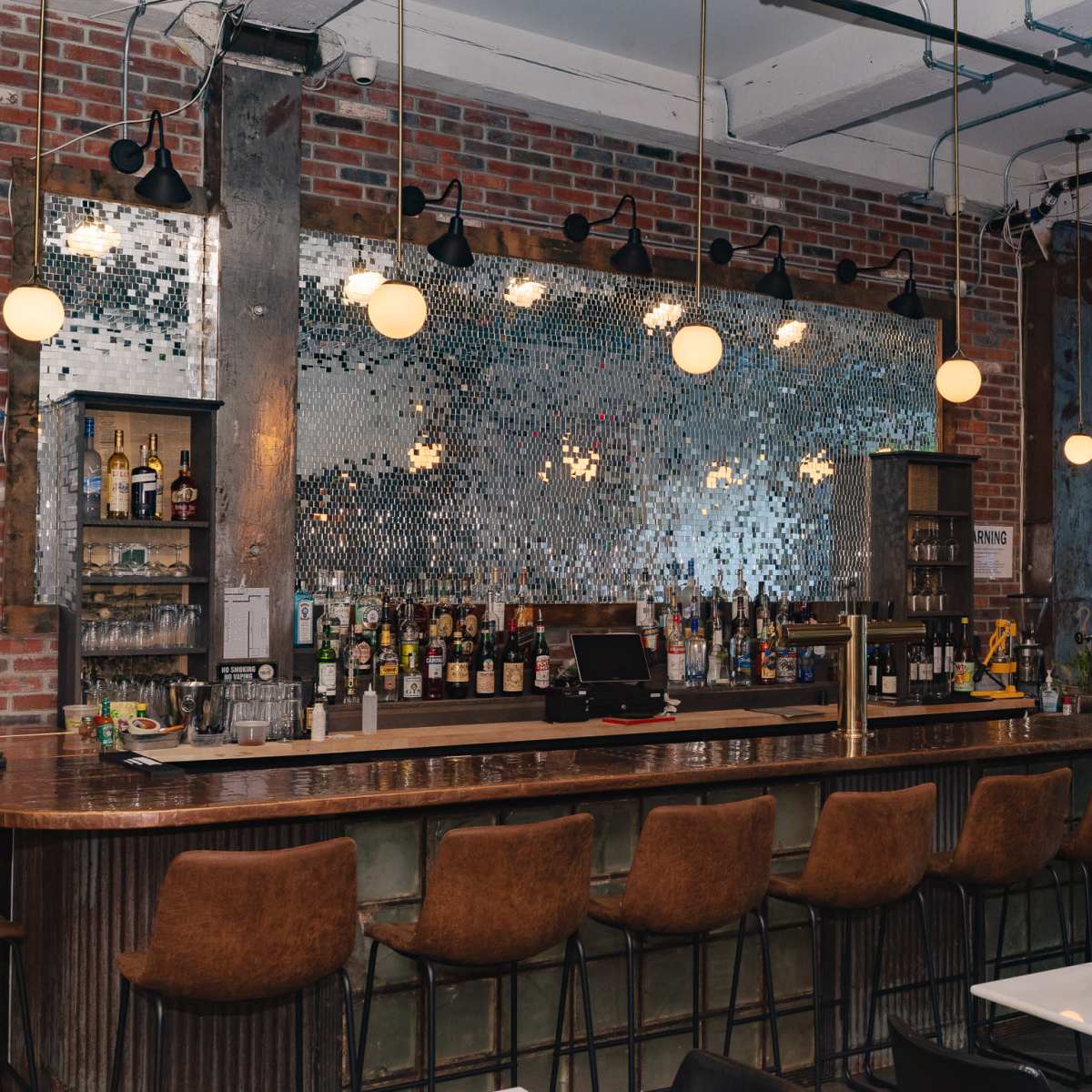A modern bar featuring a reflective mosaic back wall, a polished wooden counter, and several neatly arranged bar stools.