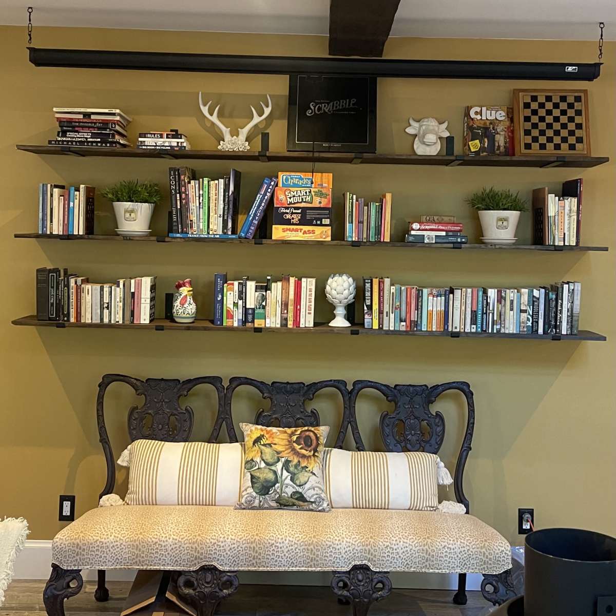 A wooden bench with patterned upholstery sits below a large, dark wooden shelf that holds an array of books and decorative items against a yellow wall.