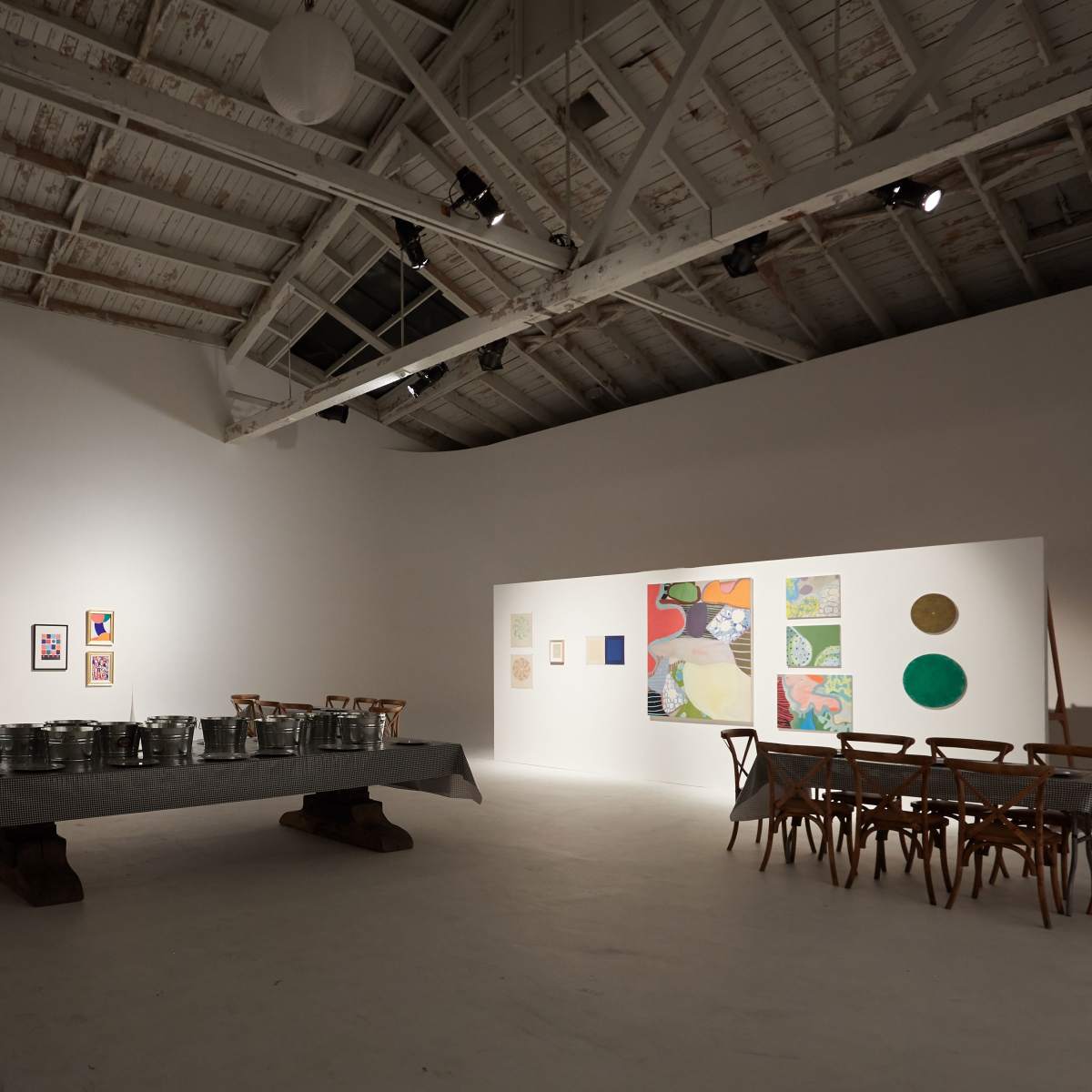 A spacious art gallery features a long table with metal containers and a wooden chair setup alongside colorful artworks on the walls.