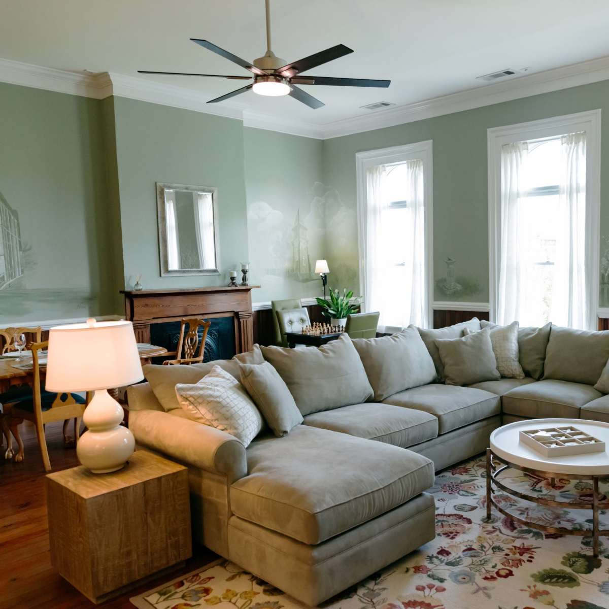 The living room features a large sectional sofa, a round coffee table, and white curtains framing the windows.
