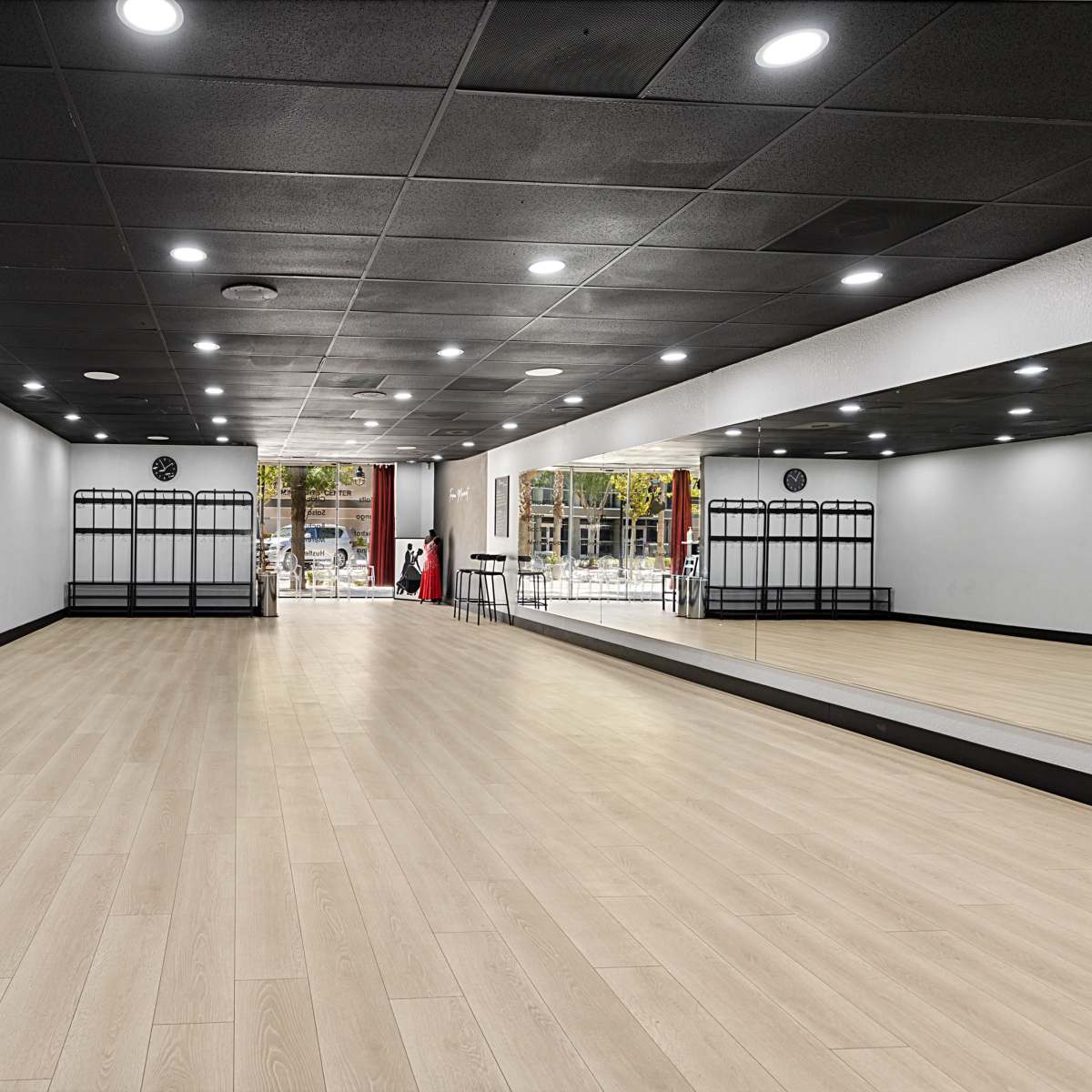 An empty dance studio with wooden flooring, mirrored walls, and a few coat racks along one side.