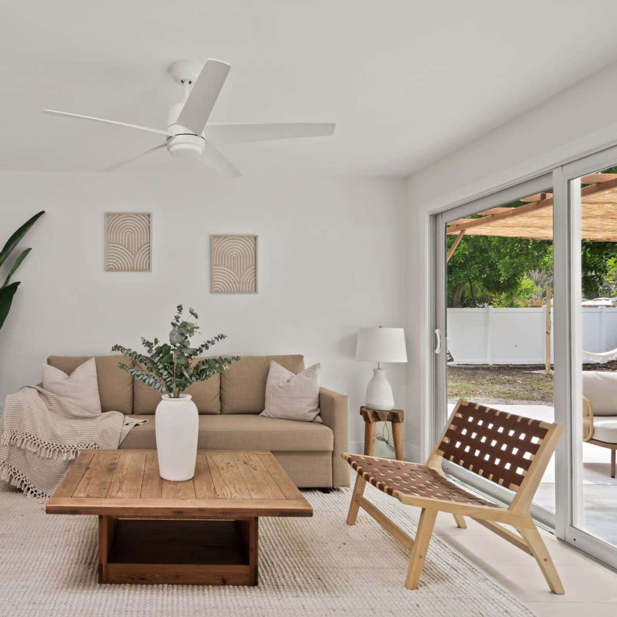 A modern living room features a beige sofa, two wooden chairs with woven seats, a wooden coffee table, and potted plants, illuminated by natural light from sliding glass doors.