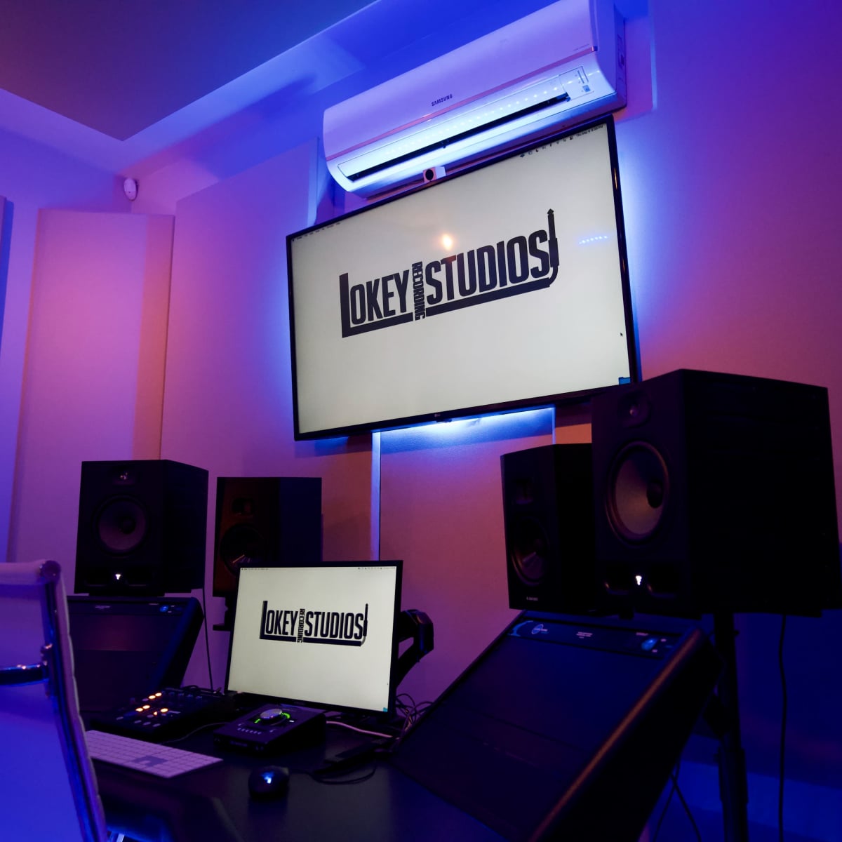 A modern recording studio with a large screen displaying "Okay Studios" and sound equipment arranged around a central desk.