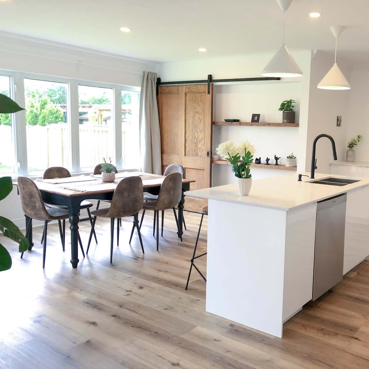 A modern kitchen and dining area featuring a central island with a sink, a wooden dining table with upholstered chairs, and large windows allowing natural light to enter.