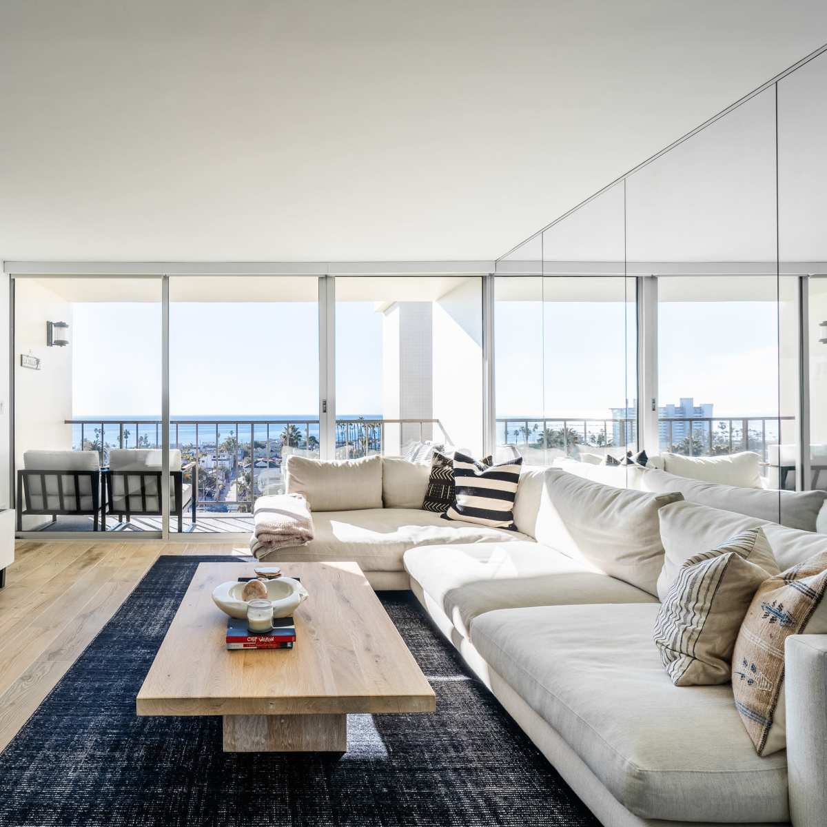 A modern living room features a large sectional sofa, a low wooden coffee table, and a wall-mounted television, with expansive glass doors offering a view of a city skyline.