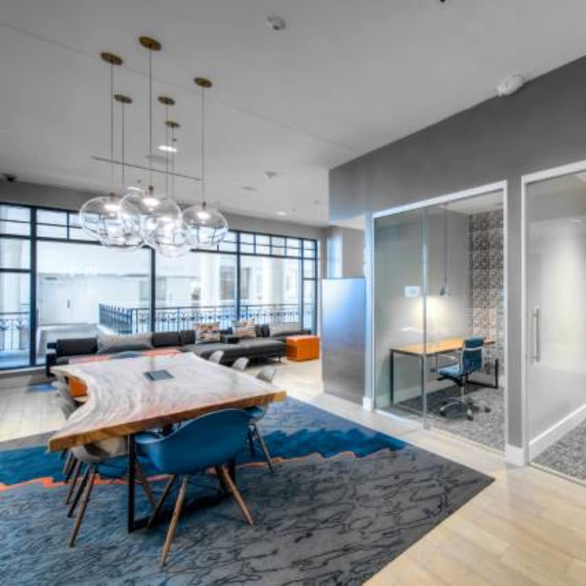 A modern office space featuring a large wooden table with blue chairs, a patterned area rug, and two glass-enclosed workstations.