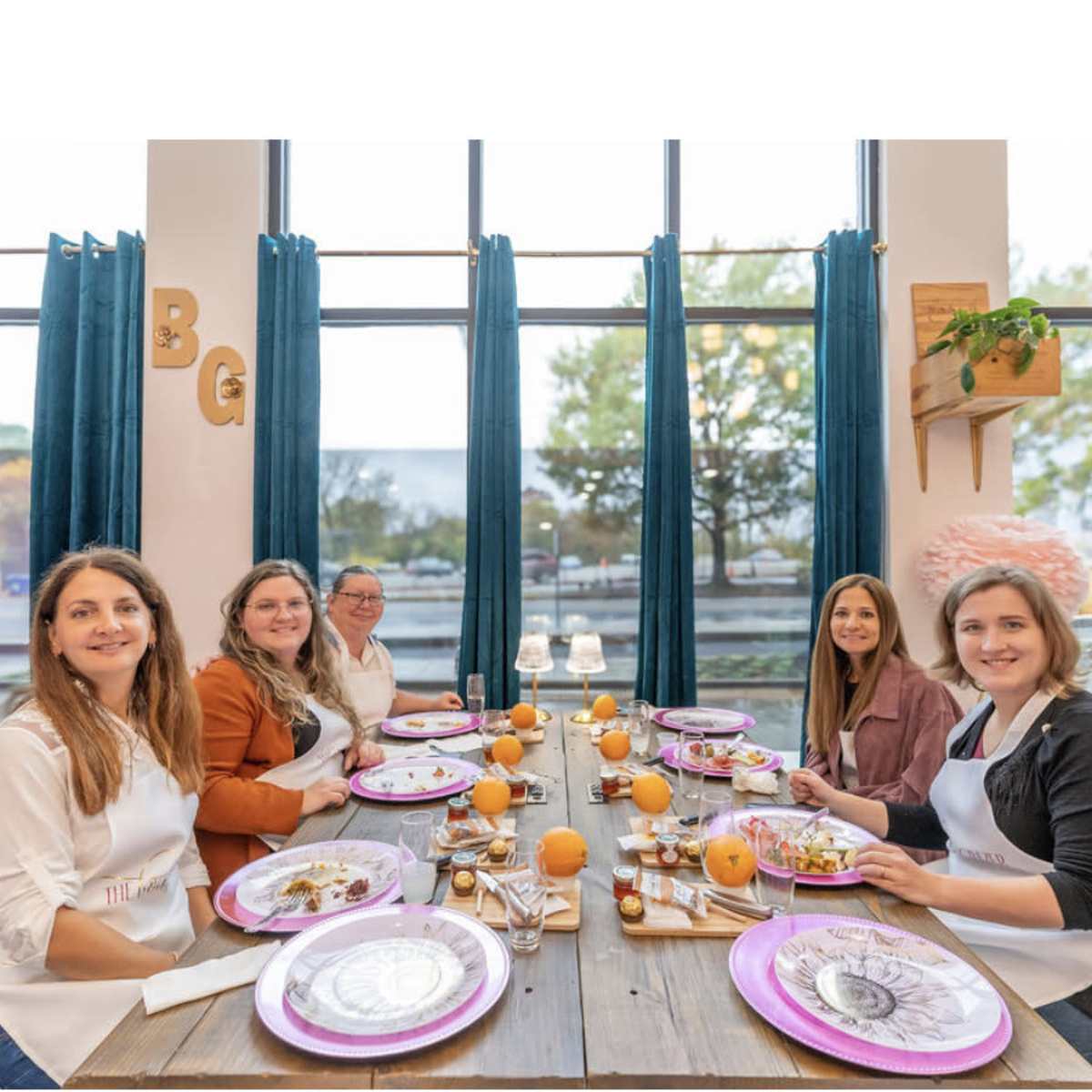 A group of five women sits at a long wooden table set with plates and food, surrounded by large windows and decorative elements.