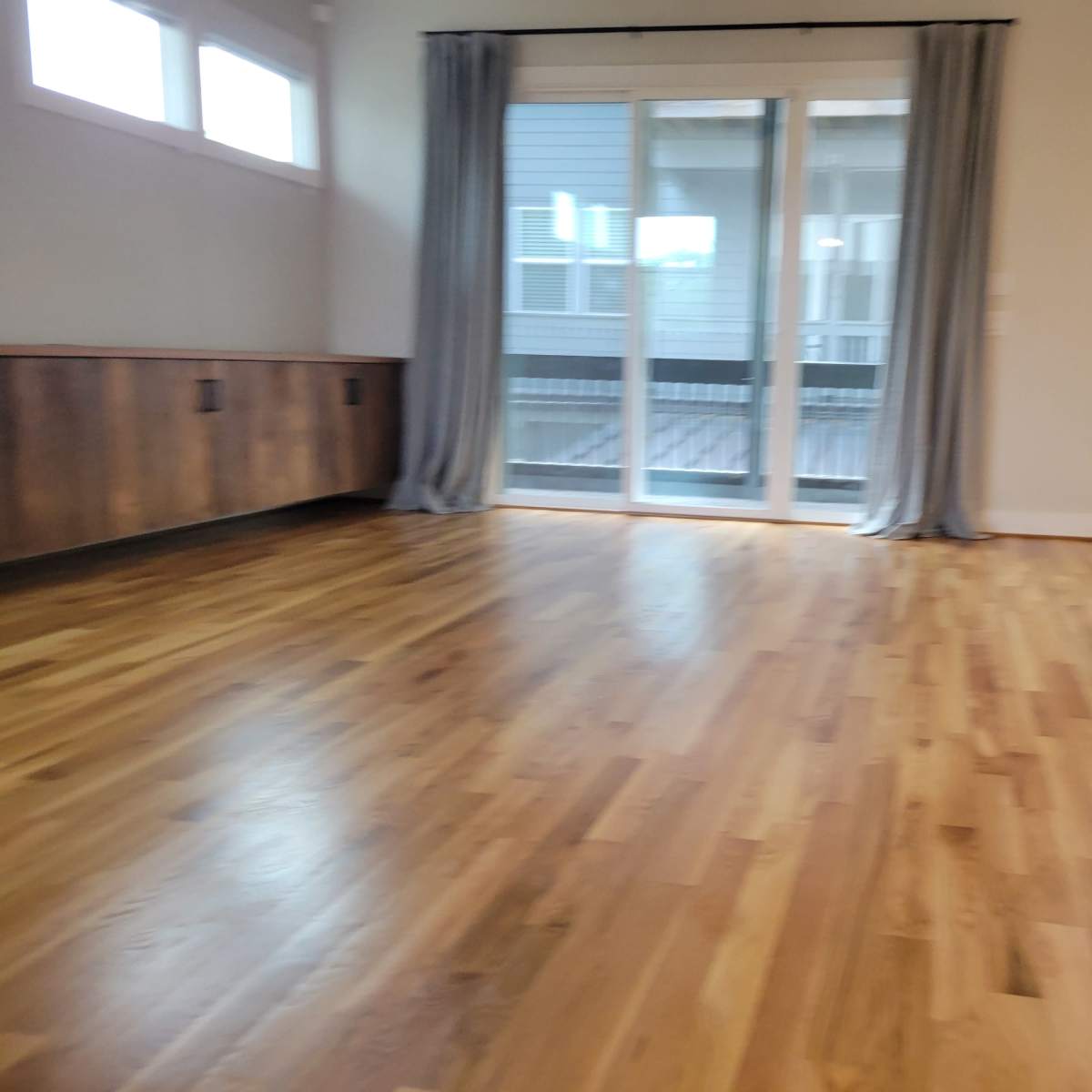A spacious room with wooden floors, large sliding doors that lead to a balcony, and gray curtains on either side.