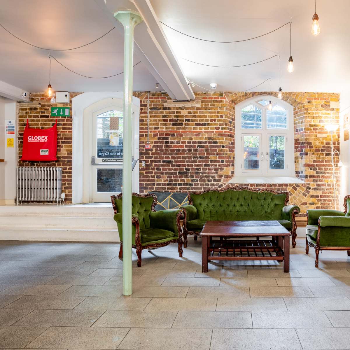 The interior features a cozy seating area with a green velvet sofa and chairs arranged around a wooden coffee table, accented by a brick wall and large windows.