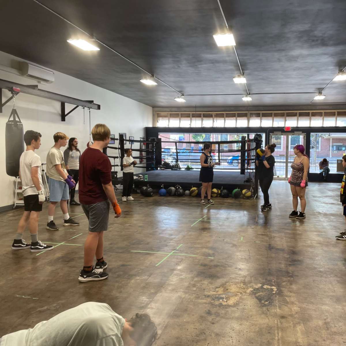 A group of young people participates in a boxing training session inside a spacious gym featuring a boxing ring and various training equipment.
