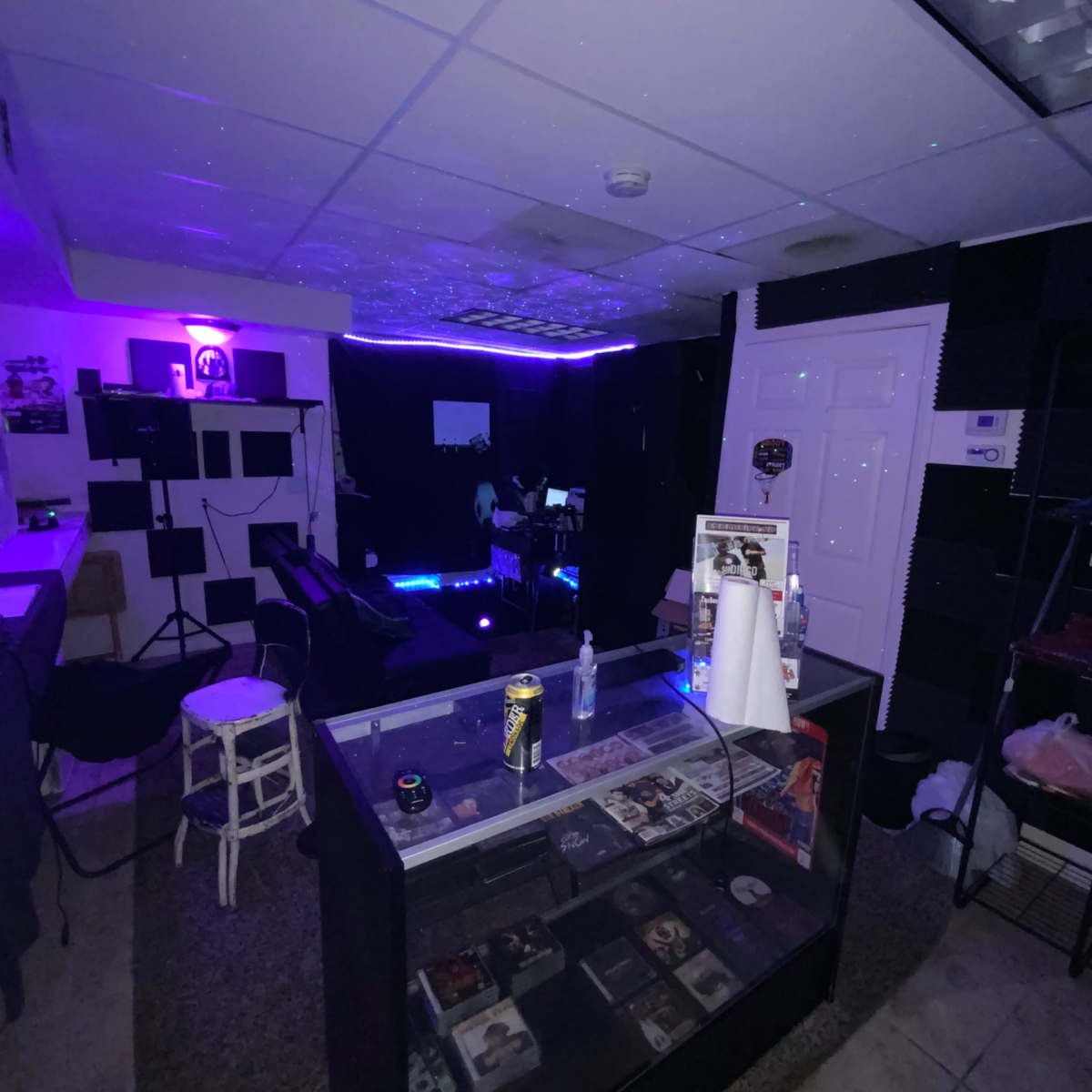 A dimly lit recording studio with purple lighting, featuring a mixing console, shelves of CDs, and soundproofing panels on the walls.
