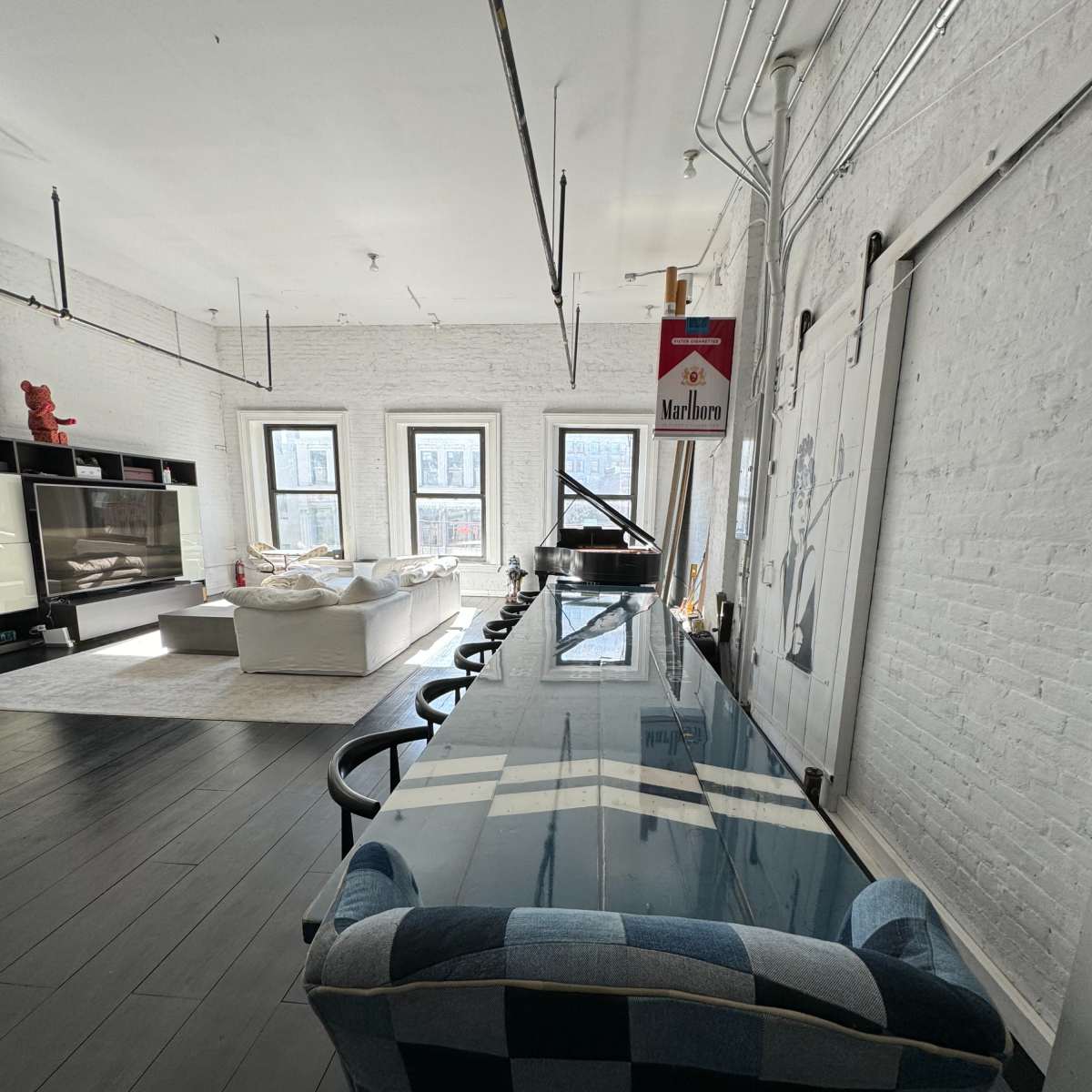 A modern, spacious dining area featuring a long glass table, a checkered chair, and a grand piano, with large windows and a white brick wall in the background.