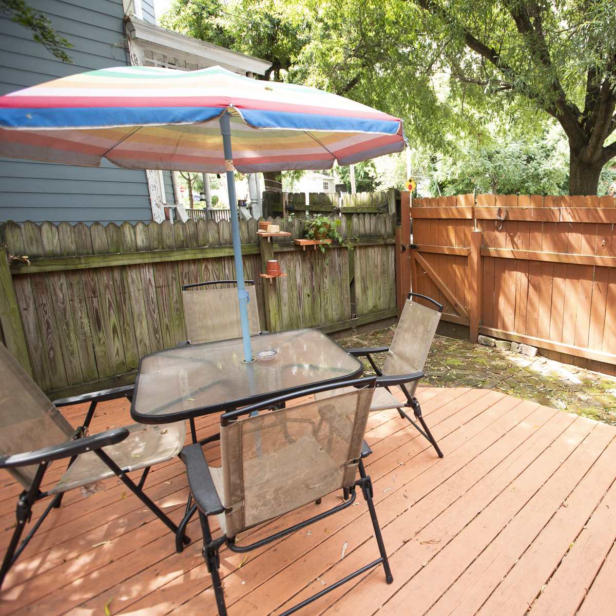 A wooden deck with a glass table and four chairs, shaded by a colorful umbrella, surrounded by a fenced yard and trees.
