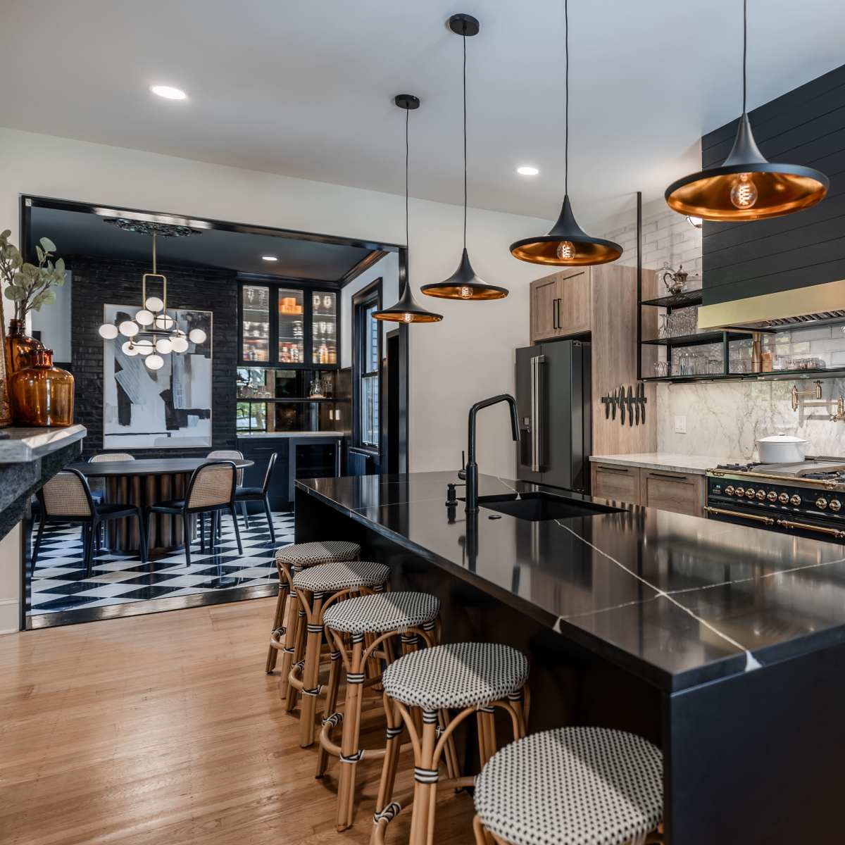 The kitchen features a central island with black countertops, pendant lighting, and wooden cabinetry, leading to a dining area with checkered flooring in the background.