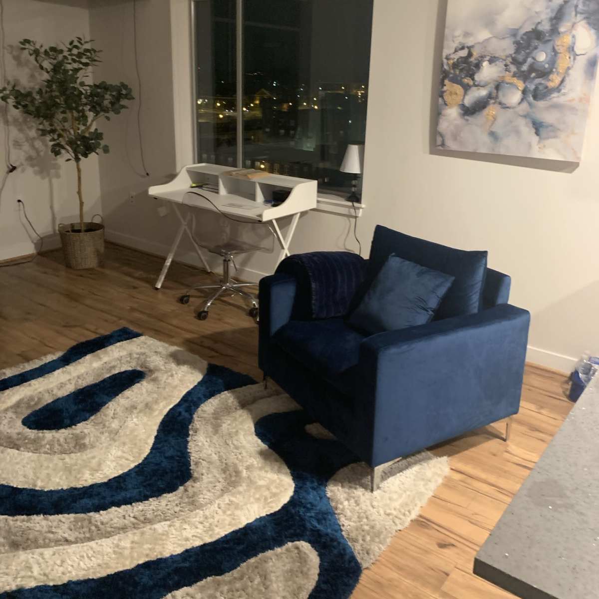 A modern apartment interior featuring a blue armchair, a white desk, and a patterned rug on a wooden floor, with a large window displaying city lights in the background.