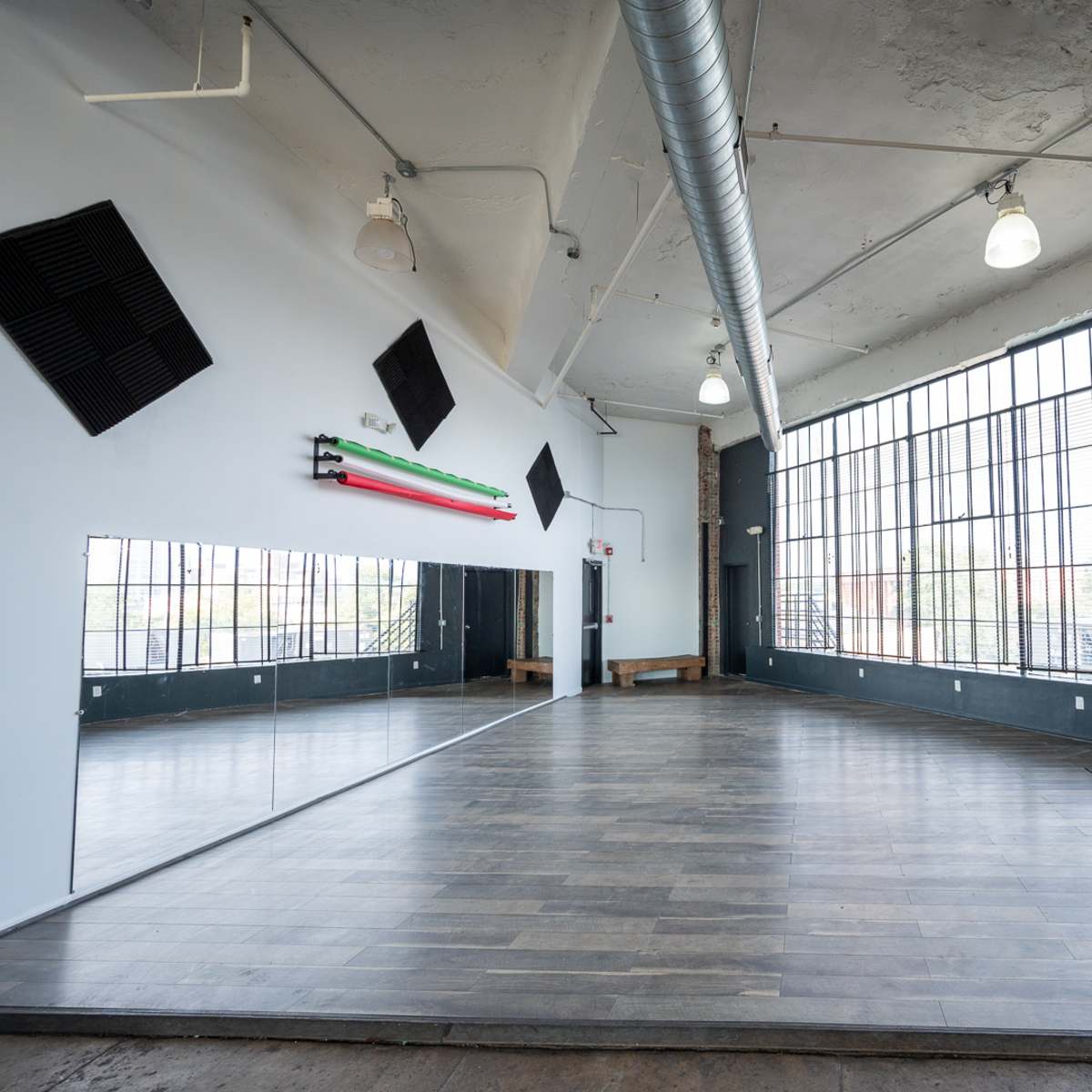 A spacious, well-lit room features large windows, wooden flooring, and a mirror along one wall.
