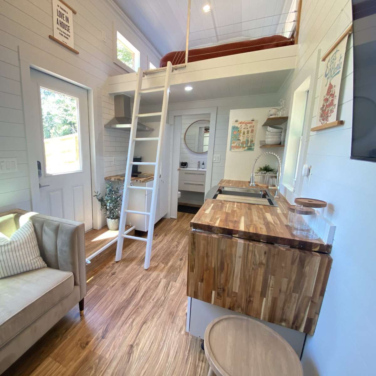 A cozy interior of a tiny house featuring a ladder to a lofted bed, a kitchenette with wooden countertops, and a living area with a beige sofa.