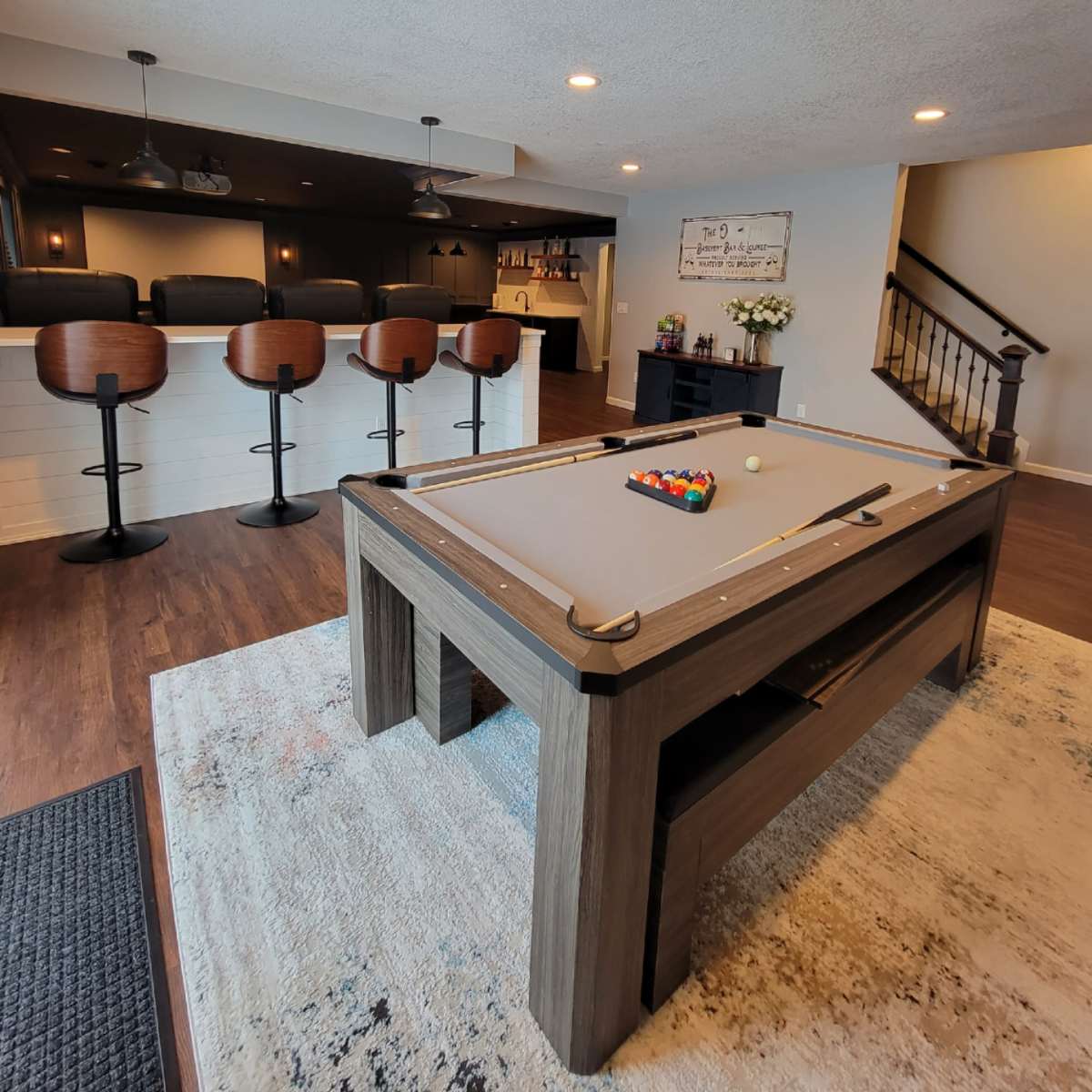 A modern game room featuring a pool table, a small bar with high stools, and a staircase leading to another level.