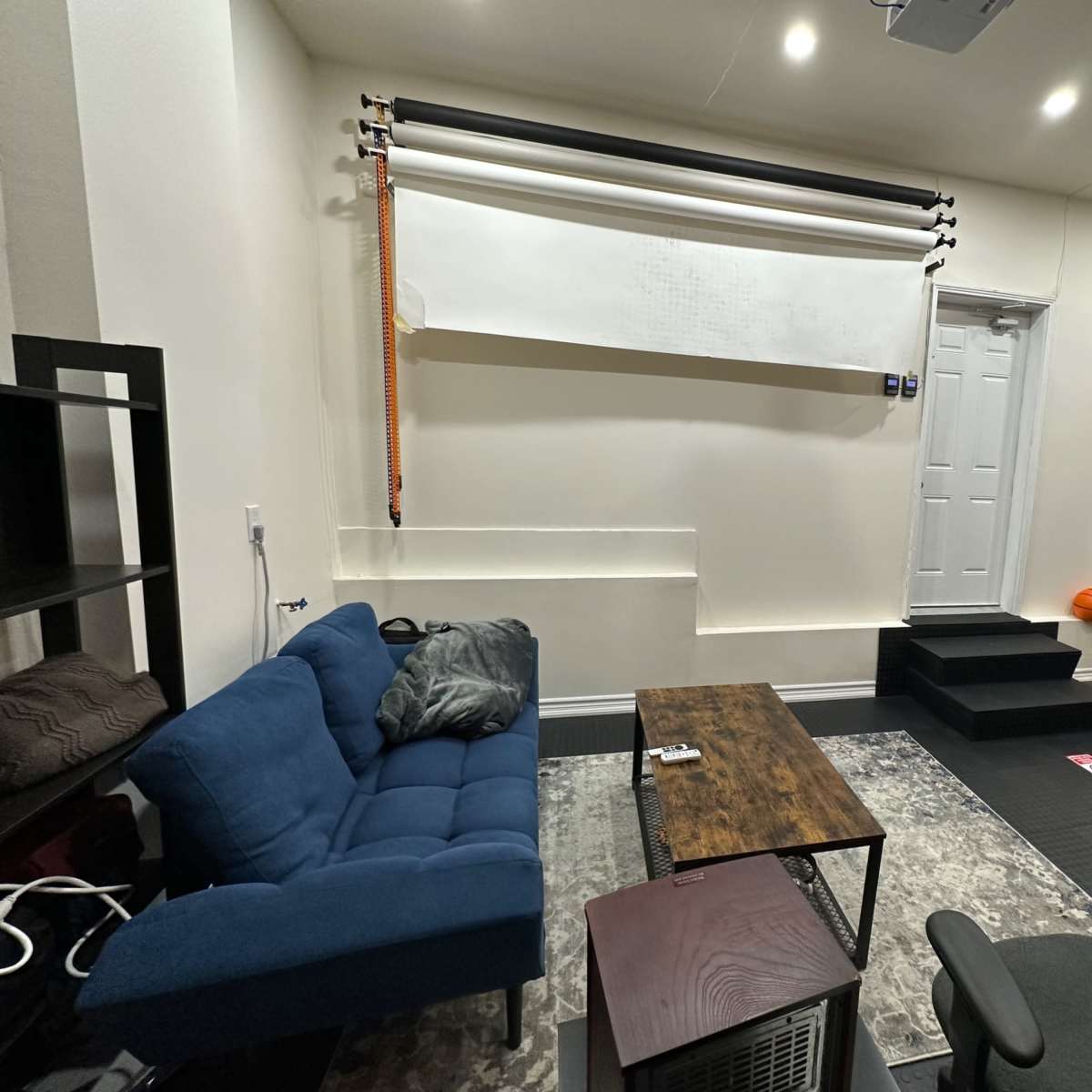 A small room with a blue couch, a wooden coffee table, and a door leading to another area, along with a whiteboard mounted on the wall and steps leading to an elevated section.