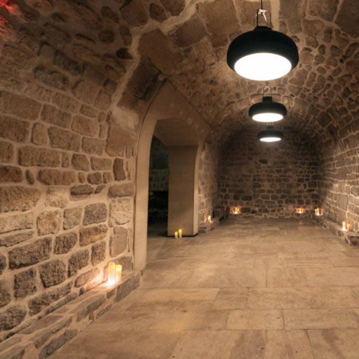 A stone-walled room with three hanging lights and candles placed along the perimeter of the floor.