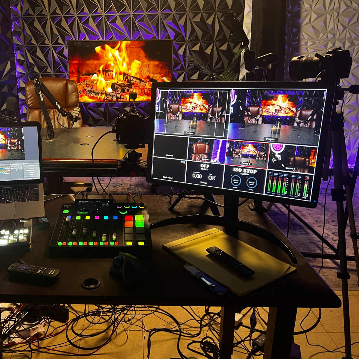 A modern video production setup with a laptop, dual monitors, a control panel, and cameras arranged around a backdrop resembling a fireplace.