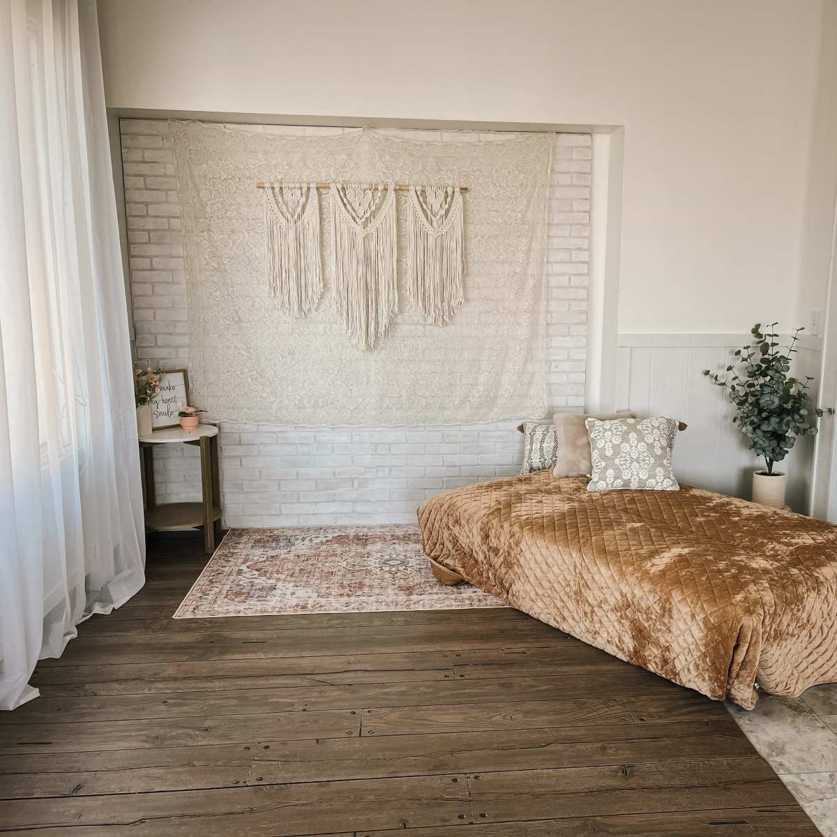 A bright, minimalist bedroom with a macramé wall hanging, a daybed covered in a textured blanket, and a small side table next to a potted plant.