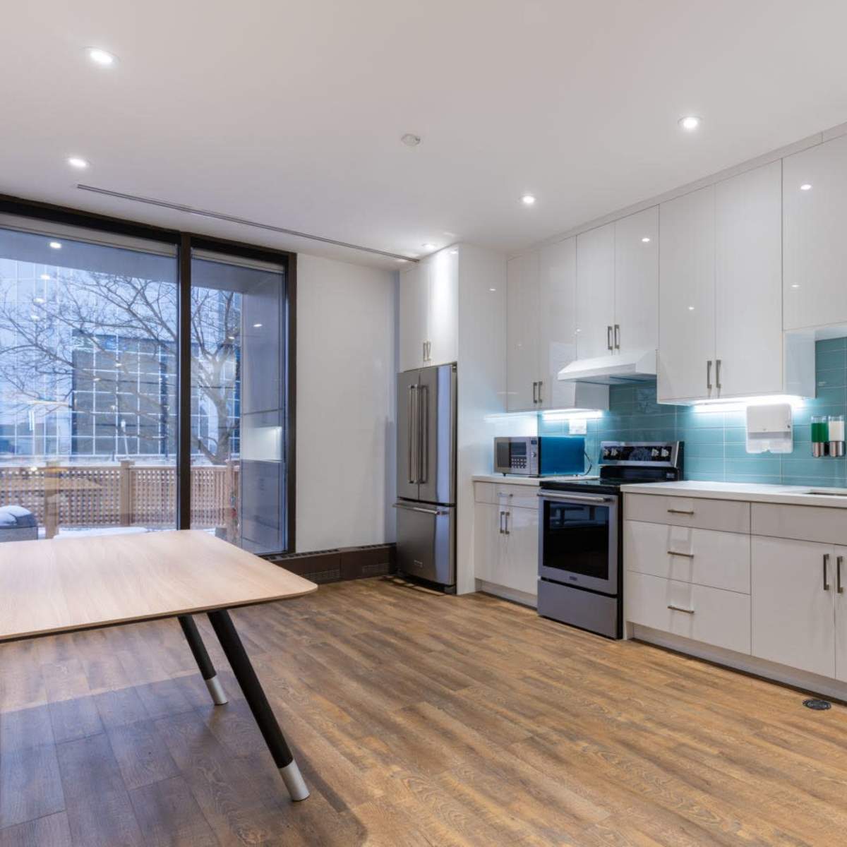 A modern kitchen features white cabinets, stainless steel appliances, and a wooden table, with a large window overlooking an outdoor area.