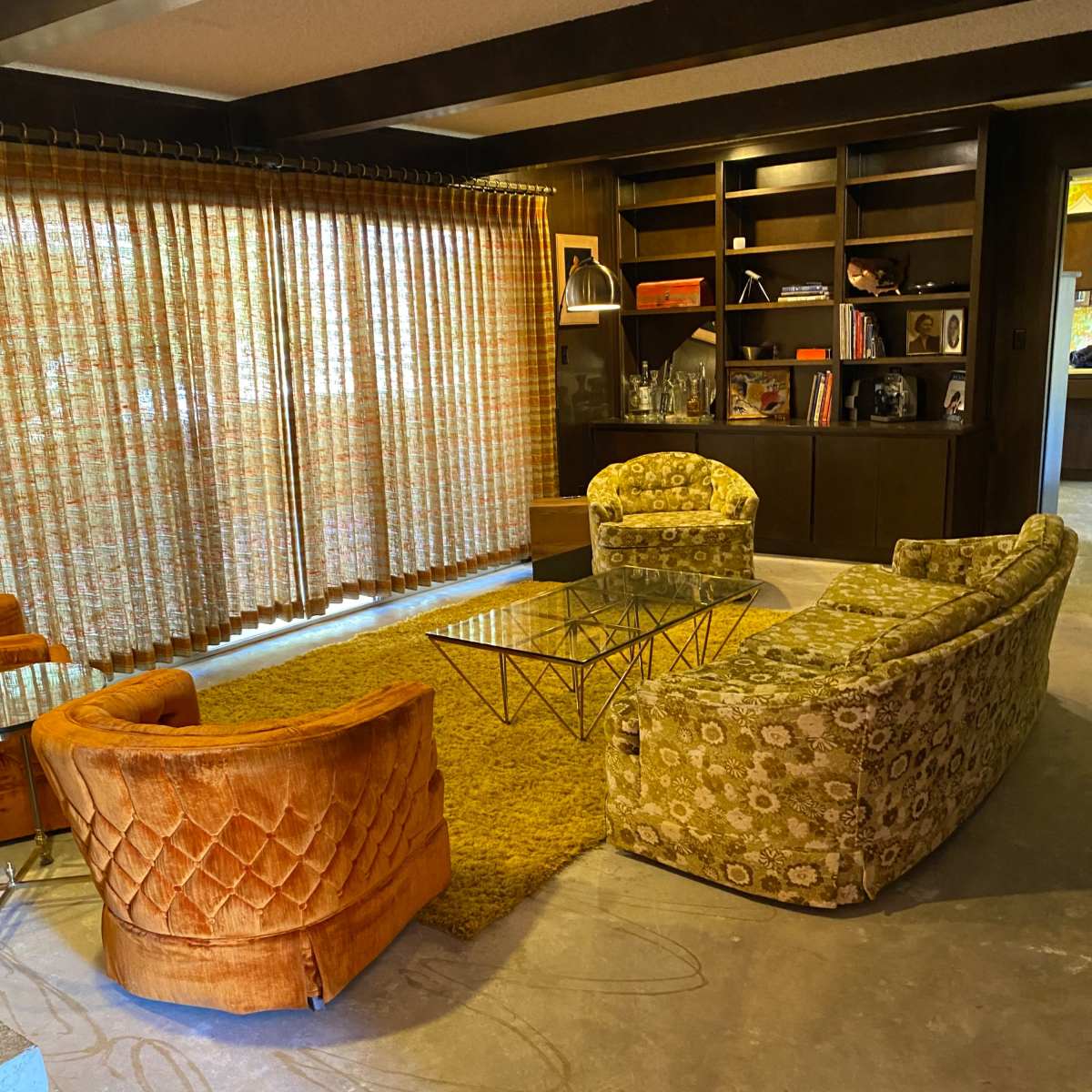 A mid-century modern living room featuring orange and green patterned sofas arranged around a glass coffee table on a textured rug, with large patterned curtains covering the windows.