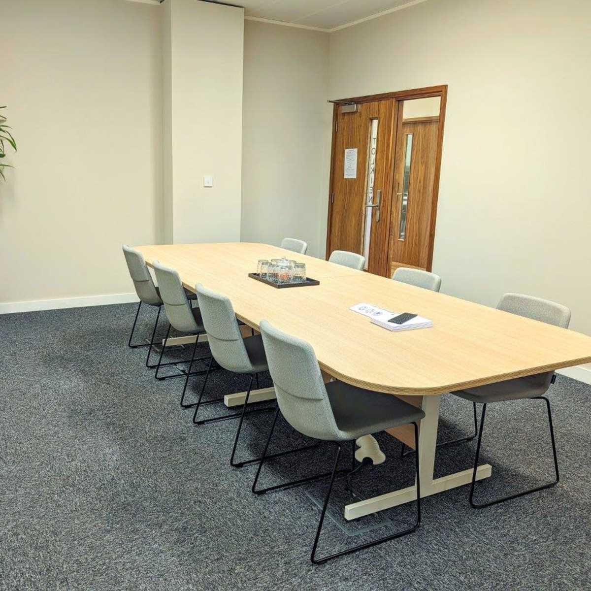 A long, rectangular conference table with eight chairs is displayed in a well-lit meeting room, featuring a potted plant in the corner and double doors leading outside.