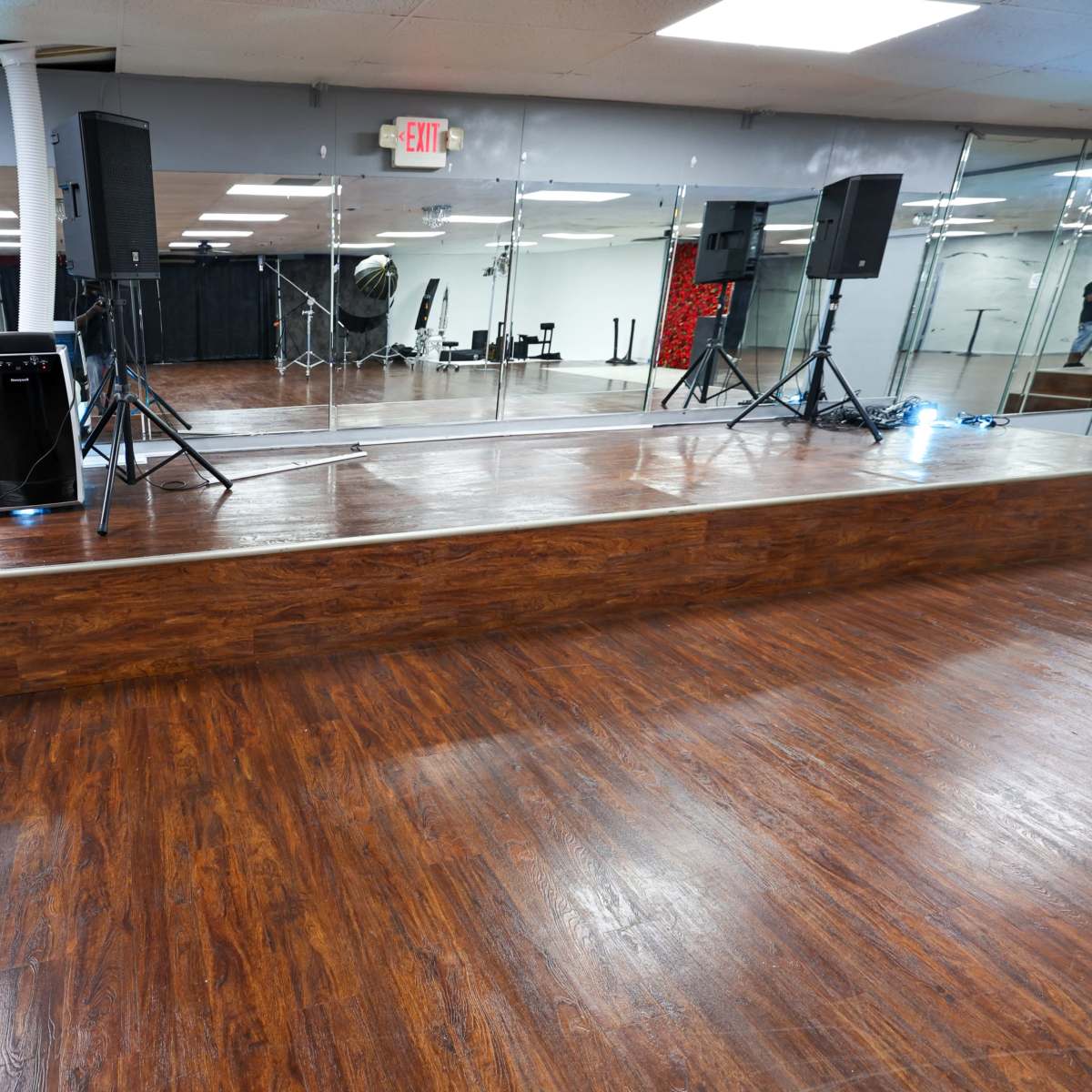 A spacious dance studio features a polished wooden floor, mirrored walls, and sound equipment set up on a raised platform.