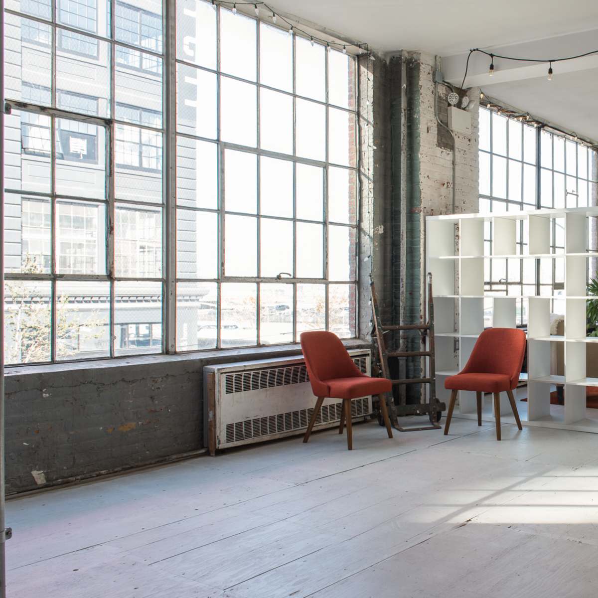 A spacious, sunlit interior with large windows, two red chairs, a white bookshelf, a gray sofa, and a potted plant on a wooden floor.