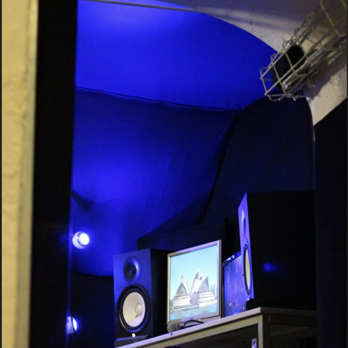 A music production studio with a computer monitor, audio speakers, and blue ambient lighting.