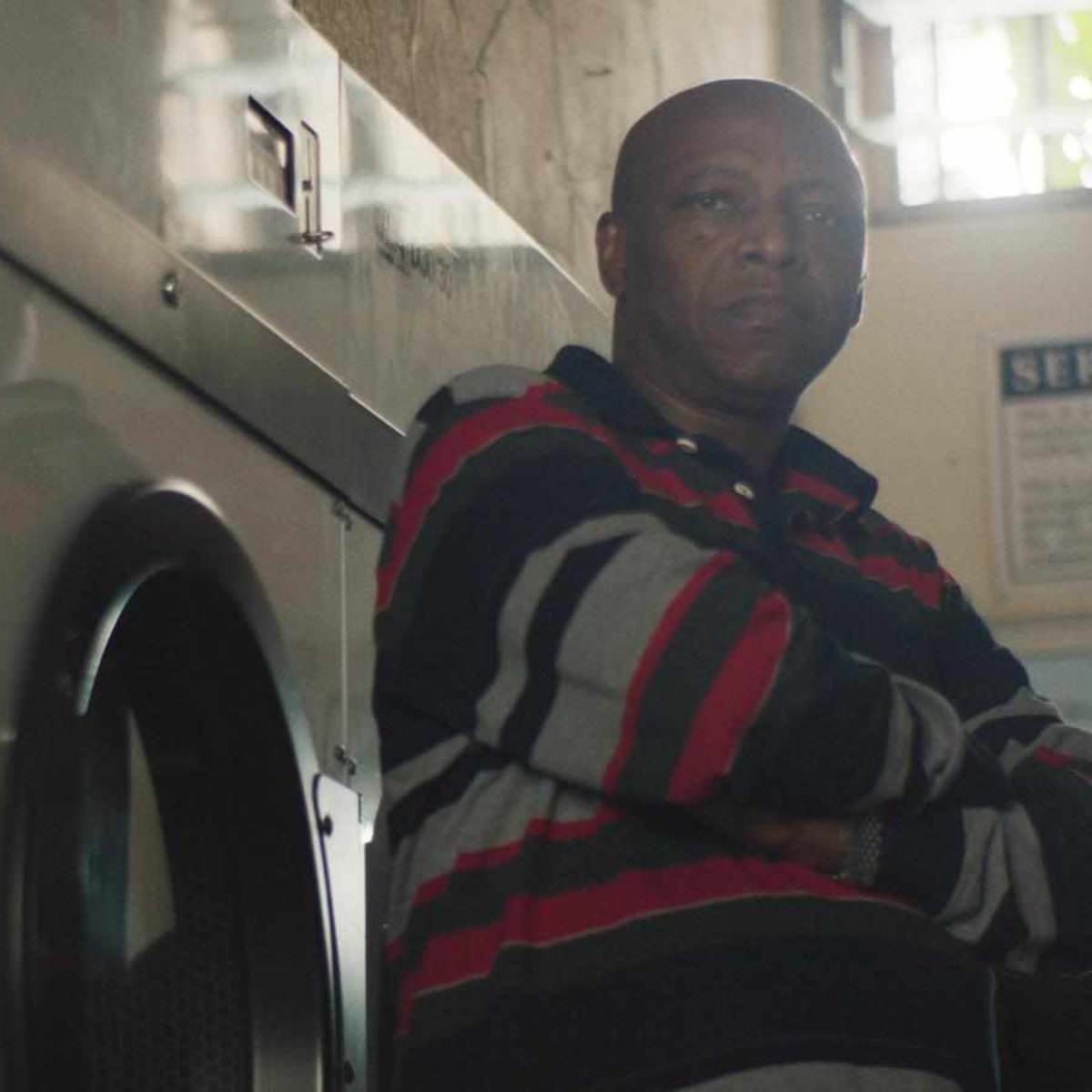 A man in a striped shirt stands with his arms crossed next to a row of washing machines in a laundry facility.