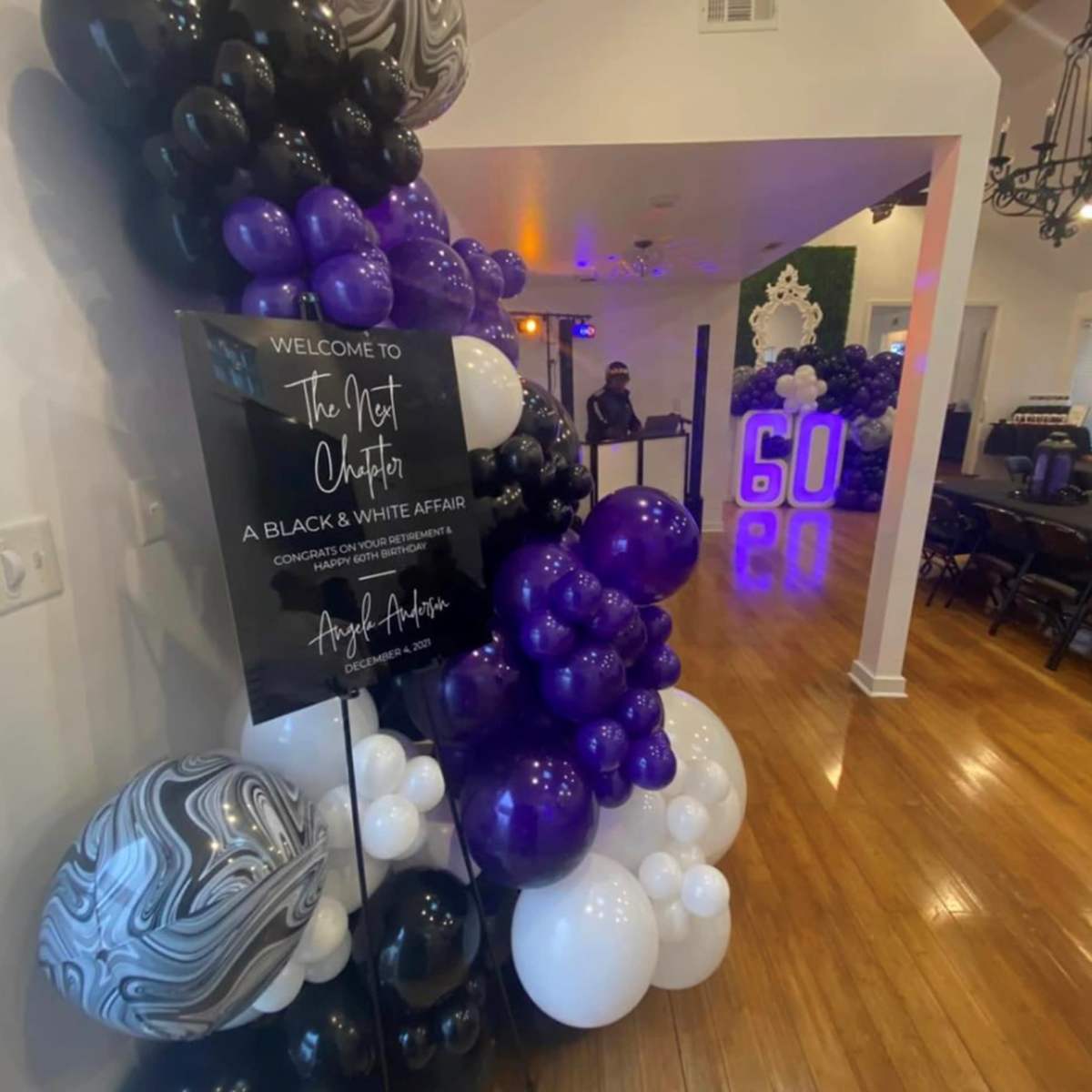 A decorated event space featuring a balloon arch in black, white, and purple, with a sign welcoming guests to a themed party.