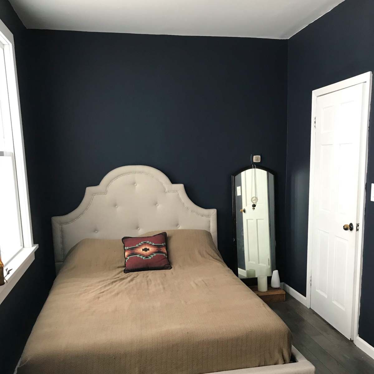 A simple bedroom with a bed featuring a tan coverlet, a patterned pillow, a full-length mirror, and dark blue walls.